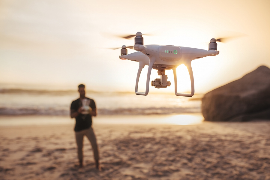 Drone being operated by a man standing in background on the beach. Young man on the sea shore flying a drone.